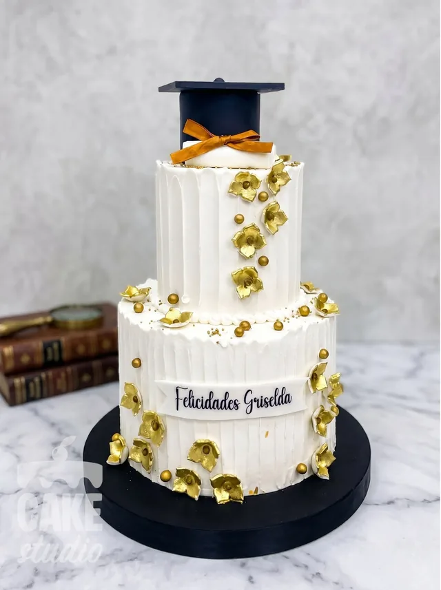 Pastel de graduación blanco con flores doradas, birrete y lazo, de Cake Studio